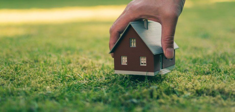 Eine Hand hält ein Modellhaus über der grünen Wiese.